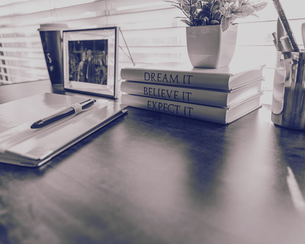 a black and white photo of books on a desk that say Dream It. Believe It. Expect It. 