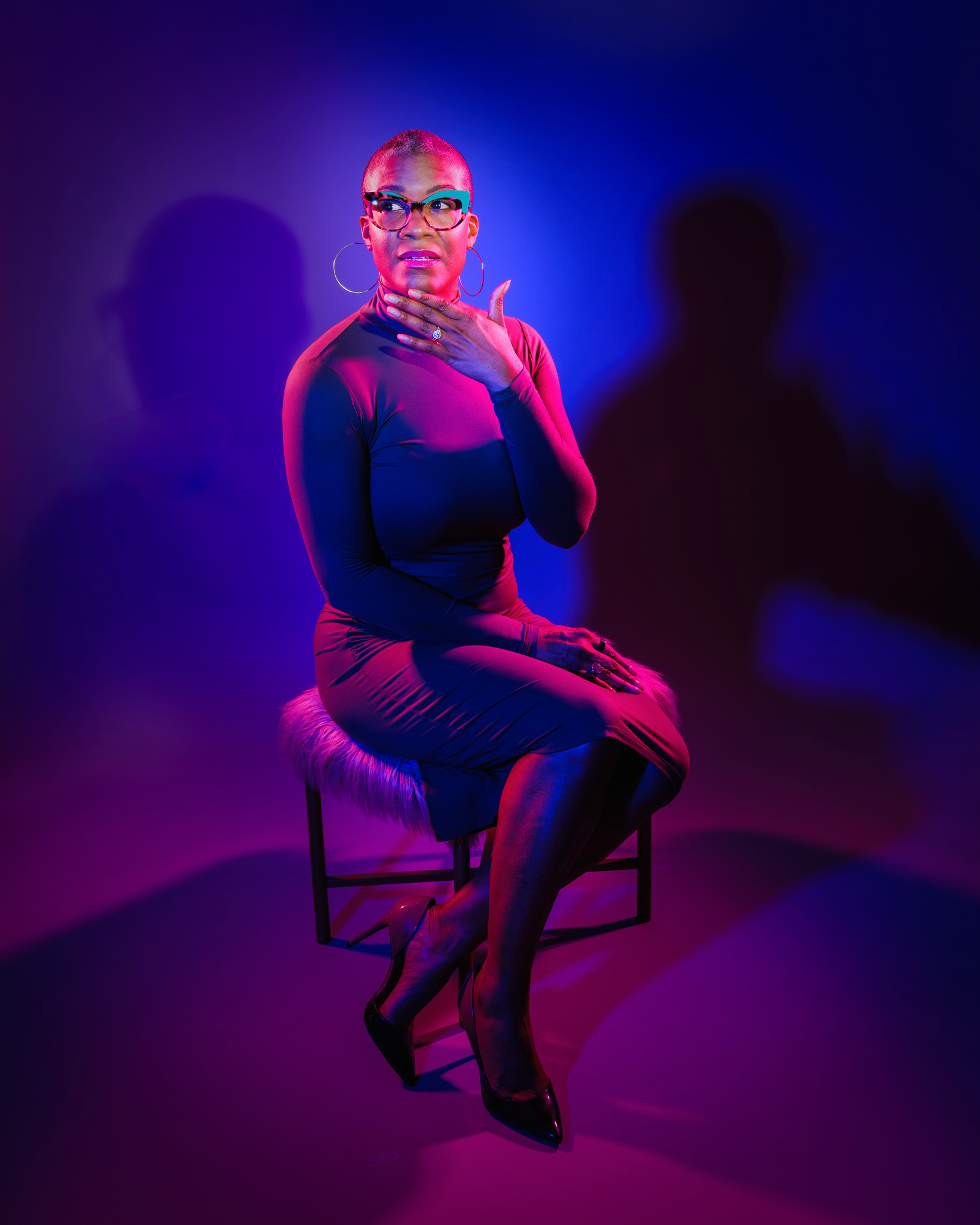 Gwendolyn "G" R Houston-Jack, creative, small biz owner, and luxury photographer sitting on a gray shaggy stool wearing an olive green mock turtleneck dress with brown ombre heels. 